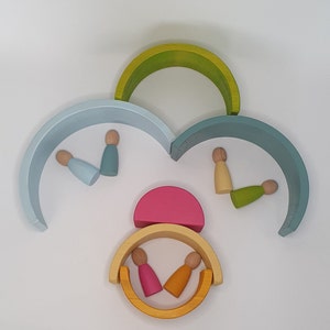 Puede incluir: Un conjunto de arcos de madera arcoíris en varios colores, incluyendo azul claro, verde, amarillo y rosa. Los arcos están dispuestos en forma de nube con pequeñas figuras de madera en su interior.