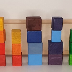 May include: A set of colorful wooden blocks stacked in different heights. The blocks are painted in shades of yellow, orange, red, brown, blue, green, and purple.