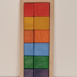 May include: A wooden box with a rainbow of colored blocks inside. The blocks are arranged in rows of two, with each row a different color. The colors are red, orange, yellow, green, blue, and purple.