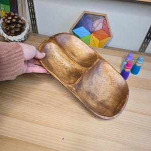 May include: A wooden serving tray with three compartments. The tray is made of a light brown wood and has a smooth, polished finish. The compartments are oval-shaped and are designed to hold different types of food or snacks. The tray is being held by a person.