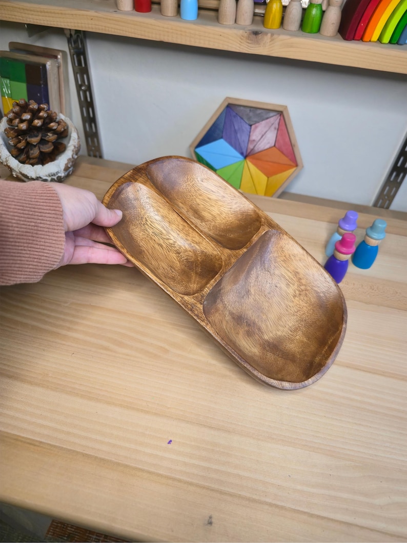 May include: A wooden serving tray with three compartments. The tray is made of a light brown wood and has a smooth, polished finish. The compartments are oval-shaped and are designed to hold different types of food or snacks. The tray is being held by a person.