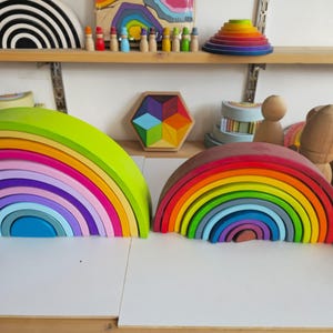May include: Two large wooden rainbow stacking toys sit side by side. The left rainbow features bright neon and pastel tones, while the right rainbow displays a classic spectrum of vibrant colors. Both are composed of multiple nested arches.