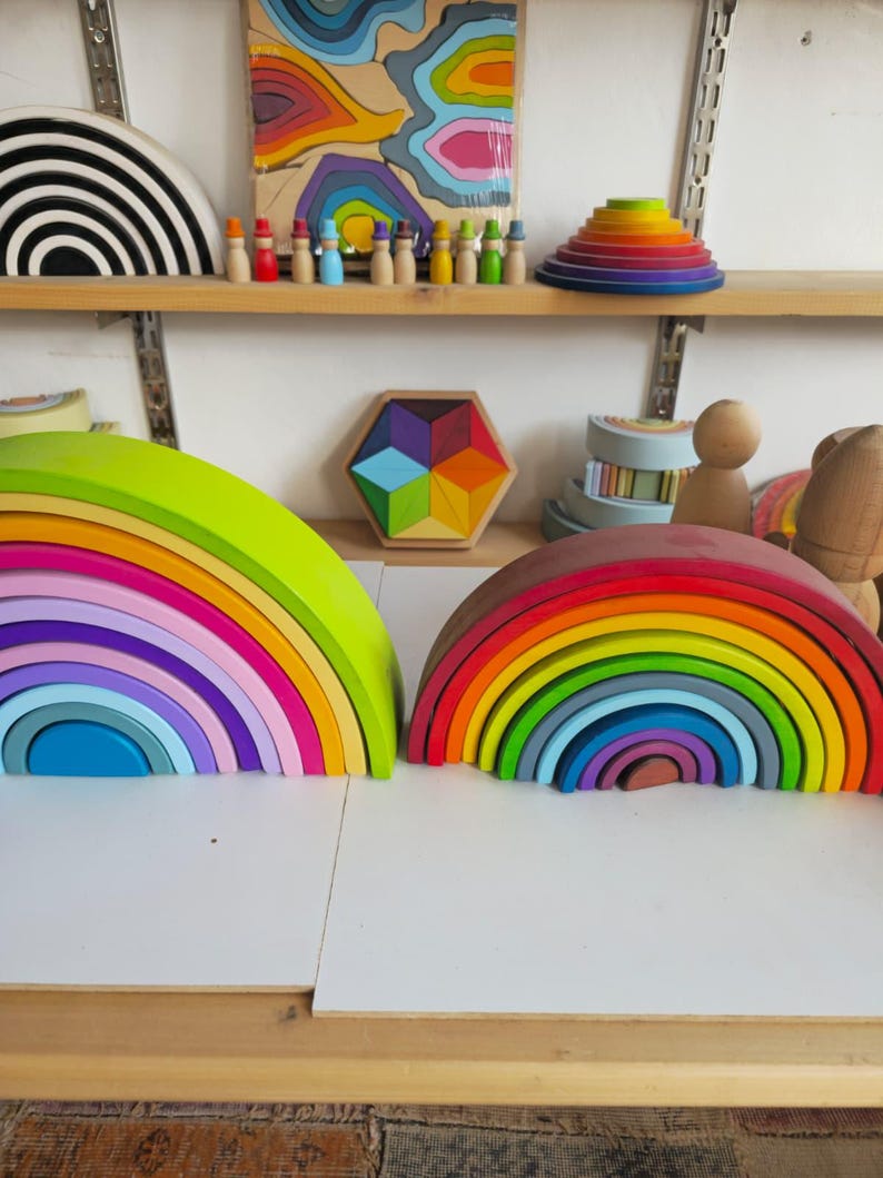 May include: Two large wooden rainbow stacking toys sit side by side. The left rainbow features bright neon and pastel tones, while the right rainbow displays a classic spectrum of vibrant colors. Both are composed of multiple nested arches.