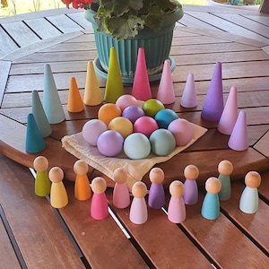 Puede incluir: Una colección de juguetes de madera que incluye varias piezas en forma de cono, bolas esféricas y muñecos de madera en un amplio espectro de colores pastel y vibrantes. Los artículos están dispuestos sobre una bolsa de tela clara, con tonos rosa, azul, amarillo, verde y morado.