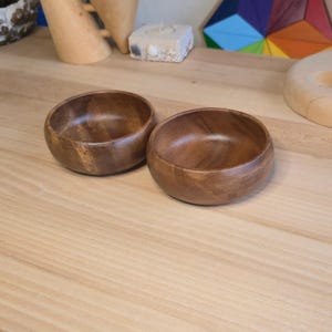 May include: Two small, round wooden bowls with a rich, dark brown color. The bowls have a smooth, polished finish and are set on a light-colored wooden surface. The bowls are likely for serving food or holding small items.