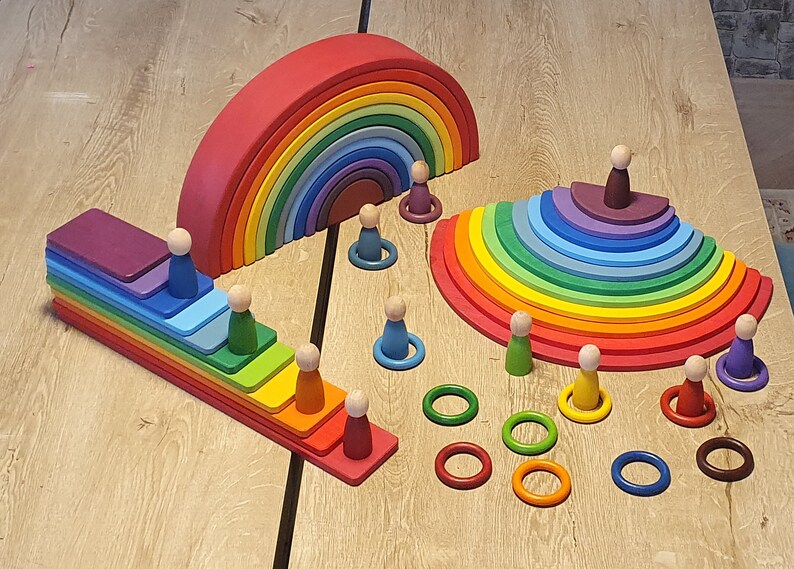 May include: A rainbow-colored wooden arch, a rainbow-colored wooden staircase, and a rainbow-colored wooden half-circle, all made of interlocking pieces. There are also small wooden figures and rings in various colors.