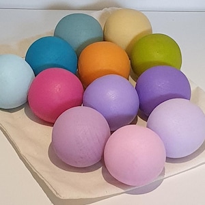 Puede incluir: Una colección de doce bolas de madera esféricas y lisas en una variedad de colores sólidos que incluyen azul claro, turquesa, rosa, naranja, amarillo, verde lima, morado y lavanda, dispuestas juntas dentro de una bolsa de algodón de color crudo.