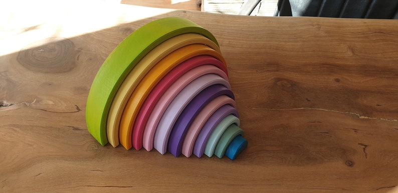 May include: A set of eleven nested wooden arches in a rainbow color gradient, ranging from lime green, yellow, and orange to various shades of pink, purple, and blue, stacked together to form a semi-circular shape on a wooden surface.