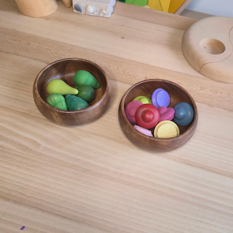 May include: Two small, round wooden bowls filled with colorful wooden toys. One bowl contains green and yellow pear-shaped toys. The other bowl holds red, pink, purple, yellow, and blue circular and heart-shaped toys. The bowls sit on a light wood surface.