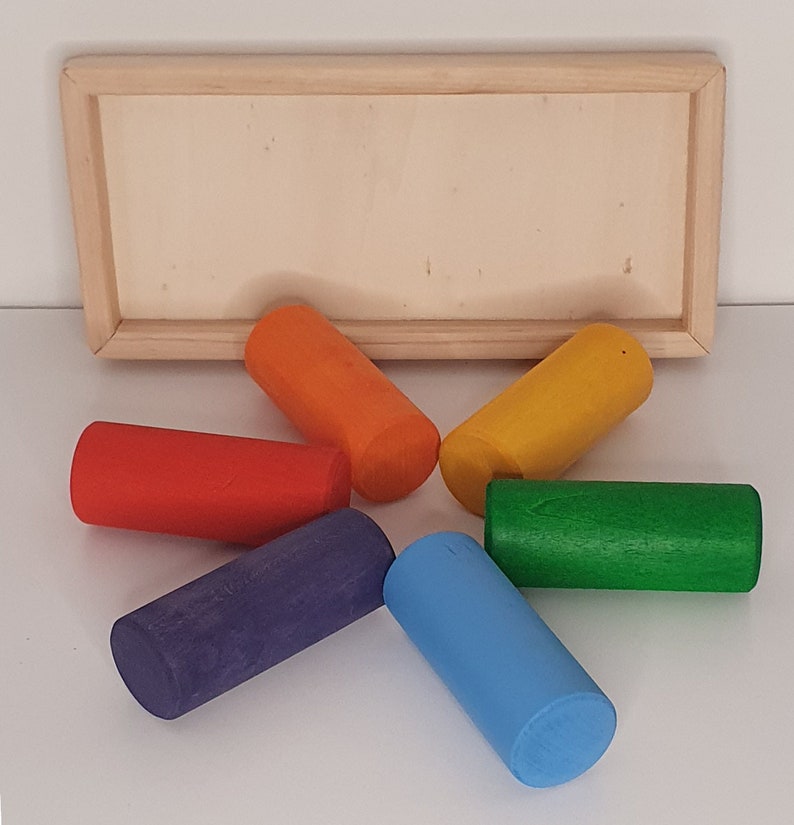 May include: Six wooden cylinders in a rainbow of colors: red, orange, yellow, green, blue, and purple. The cylinders are arranged in a star shape on a white surface.