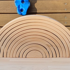 May include: A wooden rainbow arch toy made of 12 natural wood pieces. The pieces are stacked in a semi-circle shape, creating a rainbow effect.