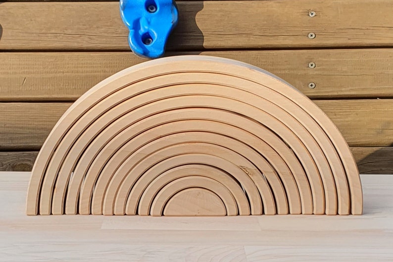 May include: A wooden rainbow arch toy made of 12 natural wood pieces. The pieces are stacked in a semi-circle shape, creating a rainbow effect.