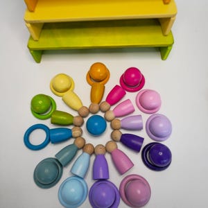 May include: A set of wooden peg dolls and matching rings arranged in a circular pattern, featuring a spectrum of colors including yellow, orange, pink, purple, blue, and green. A solid blue sphere sits in the center, with a tiered yellow and green wooden stand positioned above the arrangement.