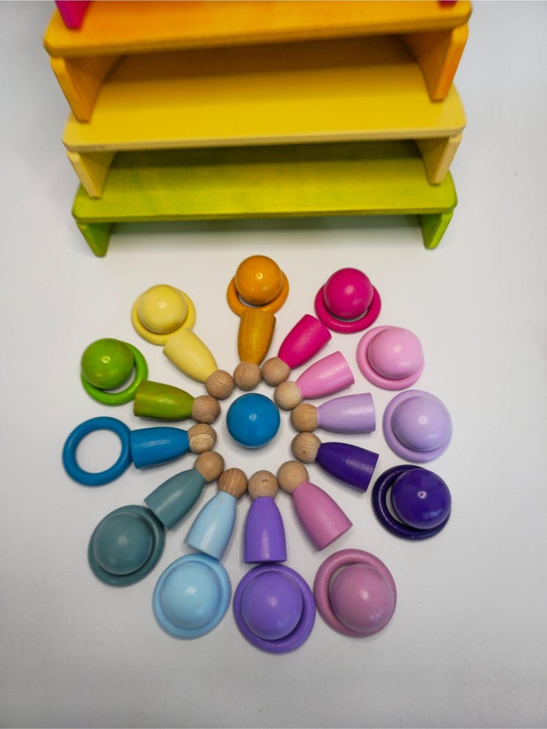 May include: A set of wooden peg dolls and matching rings arranged in a circular pattern, featuring a spectrum of colors including yellow, orange, pink, purple, blue, and green. A solid blue sphere sits in the center, with a tiered yellow and green wooden stand positioned above the arrangement.
