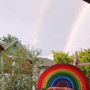 May include: A rainbow-colored wooden arch toy with 12 pieces. The toy is held in front of a house and a rainbow in the sky.