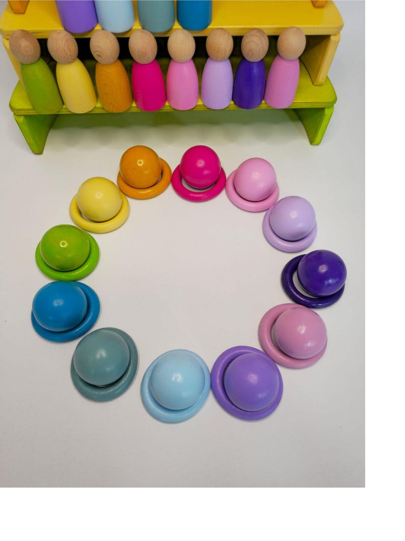 May include: A set of wooden peg dolls in various colors arranged in a row on a yellow tiered stand, with matching wooden balls and rings arranged in a circular pattern below. The colors include shades of green, yellow, orange, pink, purple, and blue.