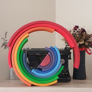May include: A rainbow-colored wooden arch made of 10 interlocking pieces. The arch is made of red, orange, yellow, green, blue, indigo, and violet pieces. The arch is sitting on a table in front of a vintage typewriter.