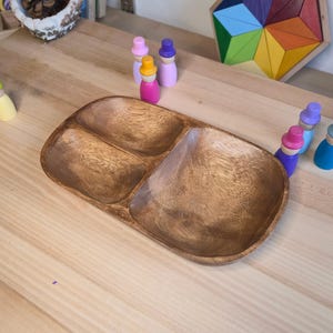 May include: A wooden serving tray with three compartments. The tray is made of light brown wood and has a smooth, polished finish. Colourful wooden peg dolls and a geometric puzzle are in the background. A pinecone is in a decorative bowl.