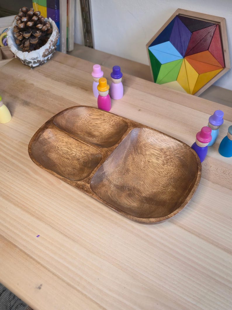May include: A wooden serving tray with three compartments. The tray is made of light brown wood and has a smooth, polished finish. Colourful wooden peg dolls and a geometric puzzle are in the background. A pinecone is in a decorative bowl.