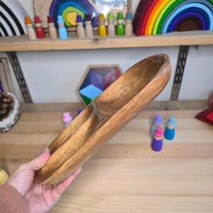 May include: A wooden serving tray with three compartments. The tray is made of light brown wood and has a smooth, polished finish. The tray is held by a person. Other wooden toys are visible in the background.