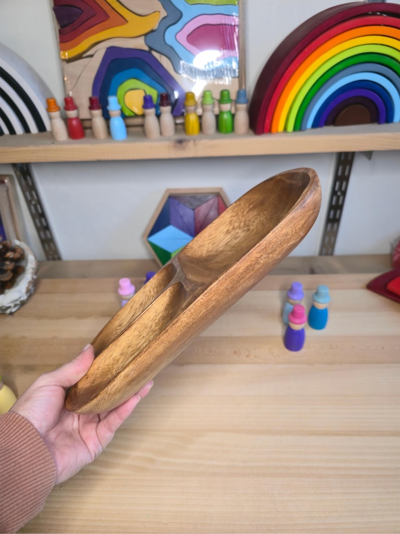 May include: A wooden serving tray with three compartments. The tray is made of light brown wood and has a smooth, polished finish. The tray is held by a person. Other wooden toys are visible in the background.