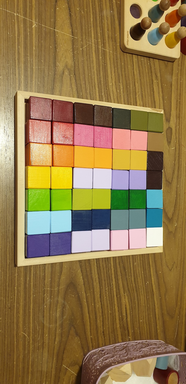 May include: A wooden educational toy featuring a grid of 64 colorful, square blocks in a rainbow of hues. The blocks are arranged in a wooden frame. Additional wooden toys are visible in the background.