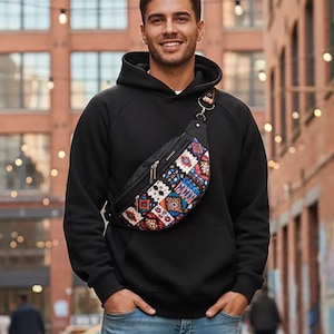 Könnte beinhalten: Ein schwarzer Kapuzenpullover, eine hellblaue Jeans mit Used-Look und schwarze Stiefel. Eine quer über der Brust getragene Gürteltasche mit einem mehrfarbigen geometrischen Muster, schwarzem Riemen und silberfarbenen Beschlägen.