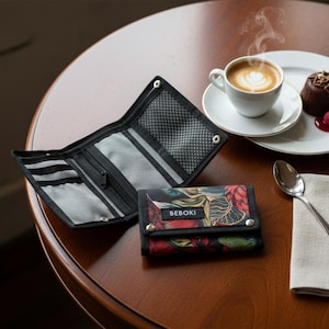 Può includere: Un portafoglio nero con stampa floreale, aperto per rivelare scomparti, è su un tavolo di legno. Un portafoglio chiuso con la scritta "BEBOKI" è accanto. Una tazza di caffè, un dolce e un cucchiaio sono anche sul tavolo.