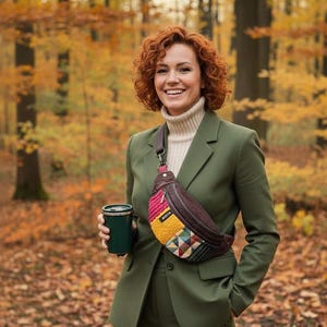 Może przedstawiać: Osoba w oliwkowym garniturze i kremowym golfie uśmiecha się, trzymając zielony kubek termiczny i nosząc brązową i patchworkową nerkę. Tło to las z jesiennymi liśćmi.