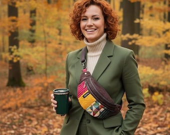 May include: A person in an olive green suit and cream turtleneck smiles, holding a green travel mug and wearing a brown and patchwork fanny pack. The background is a forest with autumn leaves.