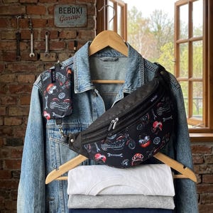 May include: A denim jacket hangs on a wooden hanger with a black fanny pack and matching small pouch, both patterned with motorcycles and tools. Below, a stack of four t-shirts in white, gray, navy, and green are visible.