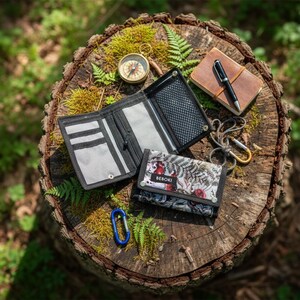 Può includere: Un assortimento di accessori per esterni, tra cui due portafogli, una bussola, un piccolo taccuino con una penna e un moschettone. I portafogli sono neri e fantasia, con il marchio "BEBOKI" visibile su uno. Gli oggetti sono disposti su un ceppo d'albero coperto di muschio.