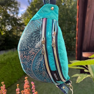 以下が含まれることがあります： ターコイズブルーと柄のファニーパック。3つのジッパー付きコンパートメントがあります。キルティングデザインと、ターコイズ、ブルー、ピンクの色合いの装飾的なマンダラ模様が特徴です。バッグはシルバーの留め金で保持され、「BEBOKI」という文字がプリントされています。