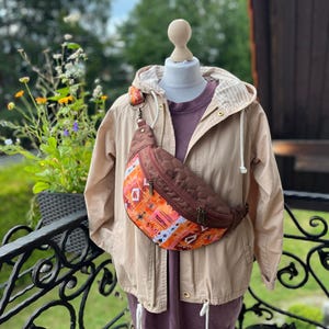 Puede incluir: Un bolso de cintura acolchado marrón con un estampado geométrico naranja y blanco se lleva sobre una chaqueta beige y una camisa morada. La chaqueta tiene capucha y cordón. El bolso de cintura tiene cremallera y correa marrones.