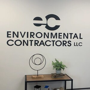 May include: A white wall with a black logo that reads "EC ENVIRONMENTAL CONTRACTORS LLC". The logo is above a black metal sculpture of three circles and a small potted plant.