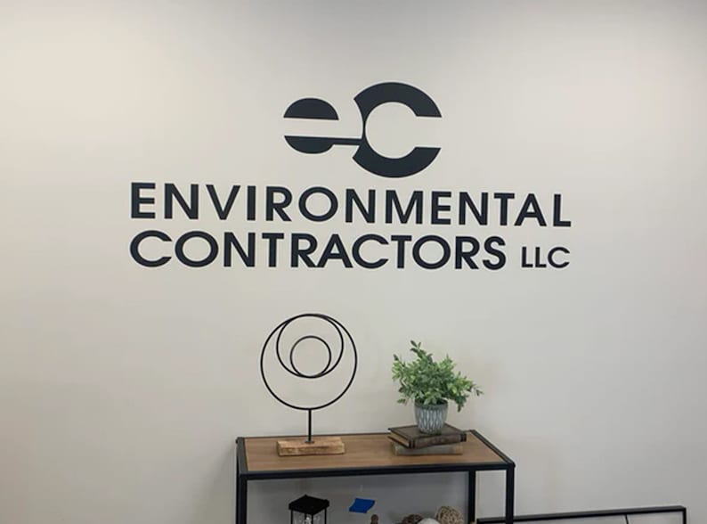 May include: A white wall with a black logo that reads "EC ENVIRONMENTAL CONTRACTORS LLC". The logo is above a black metal sculpture of three circles and a small potted plant.