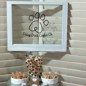 May include: A white window frame with a black paw print and the text "Daisy Doo's Cookie Co." on a glass pane. A table with a glass top and a white bowl filled with dog treats. There are three jars with lids on the table.