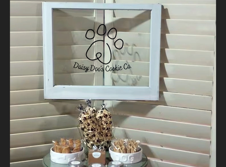 May include: A white window frame with a black paw print and the text "Daisy Doo's Cookie Co." on a glass pane. A table with a glass top and a white bowl filled with dog treats. There are three jars with lids on the table.