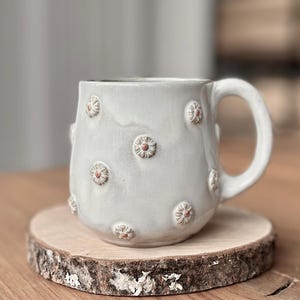 Könnte beinhalten: Hellgraue Keramik-Tasse mit einem gebogenen Henkel, verziert mit kleinen, erhabenen, gänseblümchenartigen Details mit rotbraunen Zentren. Die Tasse steht auf einem runden Holzuntersetzer. Ideal für Tee oder Kaffee.