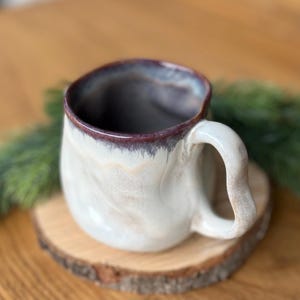 May include: A handmade ceramic mug with a unique shape and a large handle. The mug has a white and tan marbled exterior with a dark purple rim. It sits on a small wooden coaster.
