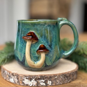 Peut inclure: Une tasse en céramique avec une finition émaillée coulante bleu sarcelle et verte. La tasse présente deux champignons sculptés en relief sur le devant, chacun avec un pied lisse de couleur claire et un chapeau brun brillant et arrondi.