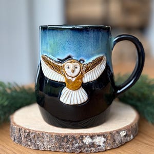 May include: A ceramic mug featuring a glossy black base that transitions into a vibrant blue glaze at the top. A sculpted, three-dimensional barn owl with outstretched wings and white and brown details is centred on the front of the mug.