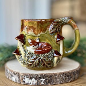 Puede incluir: Una taza de cerámica con temática de bosque. La taza presenta un zorro durmiente, setas y hojas en relieve. El asa y el borde son verdes, y el cuerpo es marrón y verde. La taza está colocada sobre una rodaja de madera.