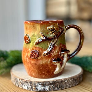 Puede incluir: Una taza de cerámica con un esmalte brillante en tonos naranja, marrón y verde. El exterior presenta decoraciones en relieve tridimensionales de una rama de árbol, varias hojas pequeñas y dos setas en el lateral junto al asa.