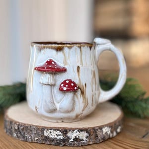Puede incluir: Taza de cerámica color crema con dos diseños de setas rojas y blancas. La taza tiene un borde y un asa marrones y está colocada sobre una rodaja de madera. La taza tiene un aspecto rústico y hecho a mano.