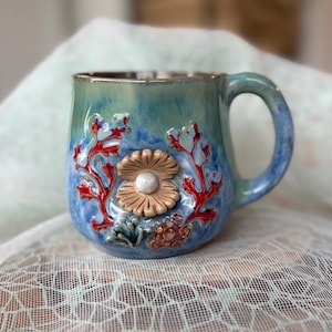 Puede incluir: Una taza de cerámica con esmalte degradado en azul y verde que presenta un relieve tridimensional de una almeja beige abierta con una perla blanca, rodeada de ramas de coral rojo esculpidas y pequeños elementos marinos texturizados.