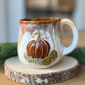 Peut inclure: Une tasse en céramique avec une base blanche brillante et un vernis coulant marron sur le bord. L'avant présente une citrouille orange foncé en relief tridimensionnel avec des feuilles vertes et des vignes texturées sur la poterie.