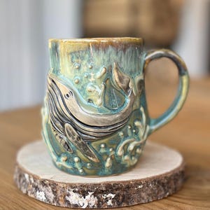 Puede incluir: Taza de cerámica turquesa y marrón con un diseño de ballena esculpida. La taza está sobre un posavasos de madera redondo. Los detalles de la ballena y el agua son en relieve.