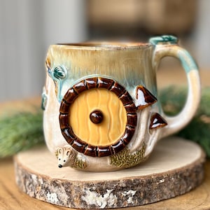 Könnte beinhalten: Eine Keramiktasse mit einem skurrilen Design, das eine gelbe Tür mit braunem Rahmen, Pilzen und einem kleinen Igel zeigt. Die Tasse hat eine beige und blaue Glasur und steht auf einem Holzuntersetzer.