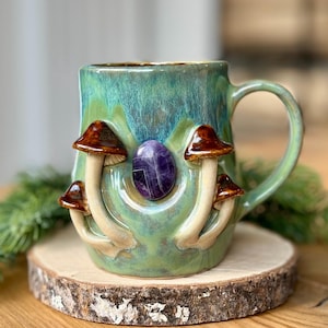 May include: A ceramic mug with a teal and green glaze, adorned with four mushroom figures and a purple gemstone. The mug sits on a wooden coaster, with a sprig of greenery in the background. The mug has a handle.